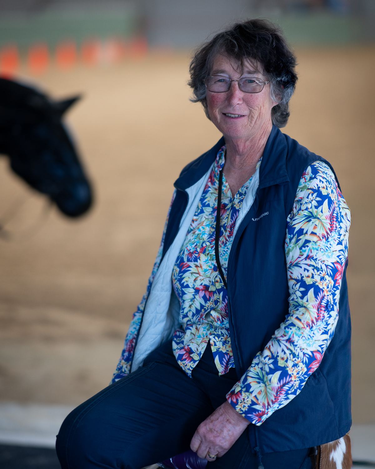 A woman with short grey hair in a floral shirt and navy vest, seated in the arena
