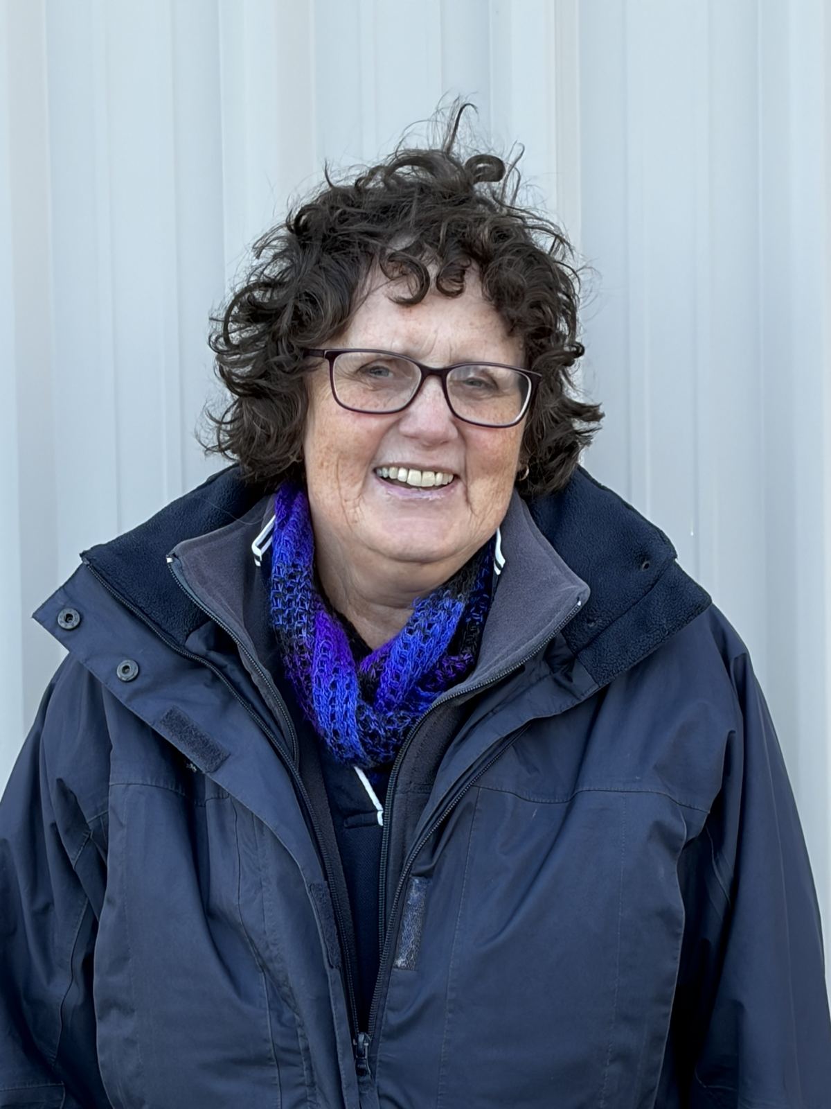 A smiling woman with curly hair and glasses, wearing a grey jacket