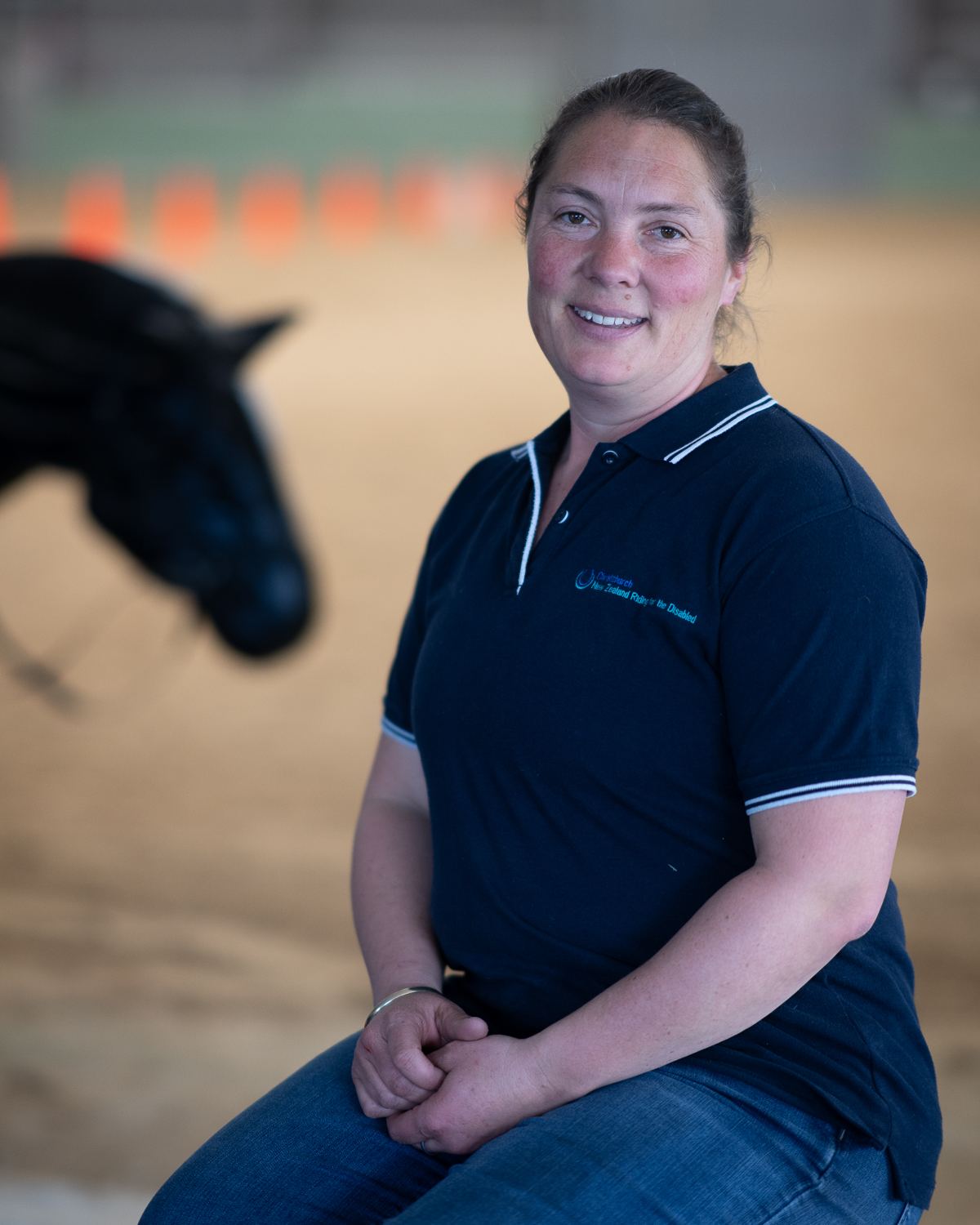 A smiling woman with her hair tied back, in a navy polo shirt, seated in the arena