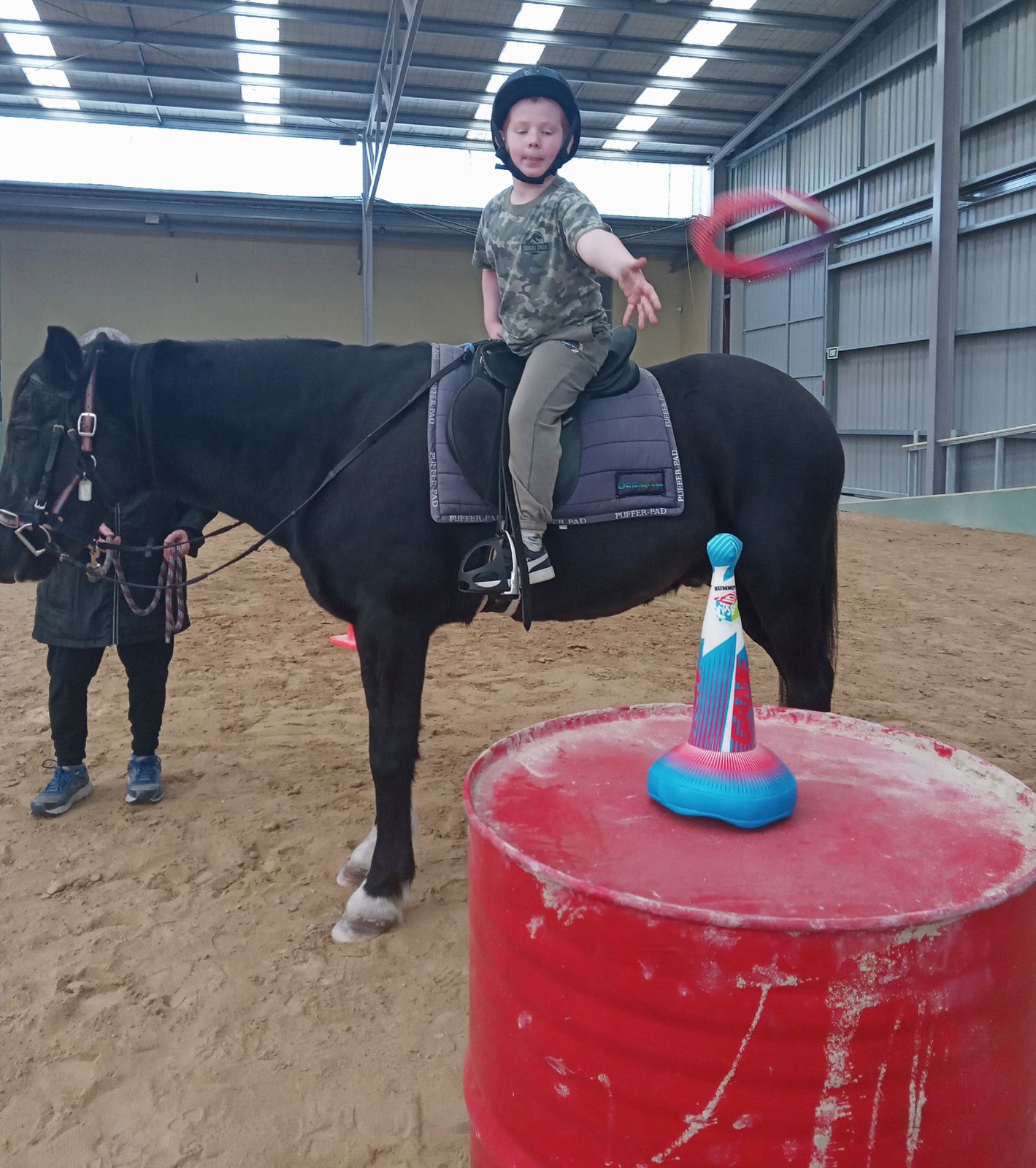 A rider on a black horse in the arena reaching towards a red barrel