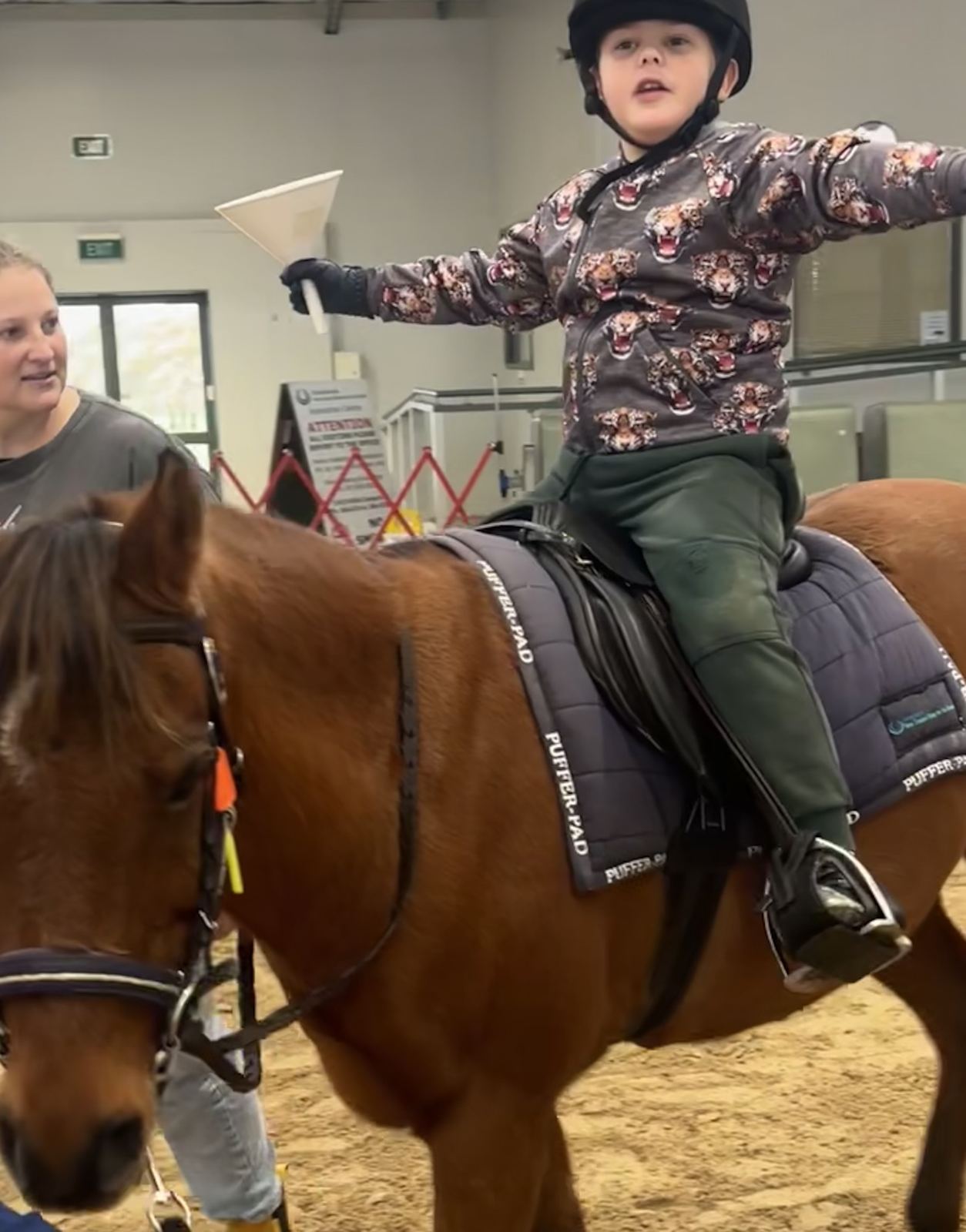 A grinning young rider stretching his arms out wide while sitting on a bay horse in the arena