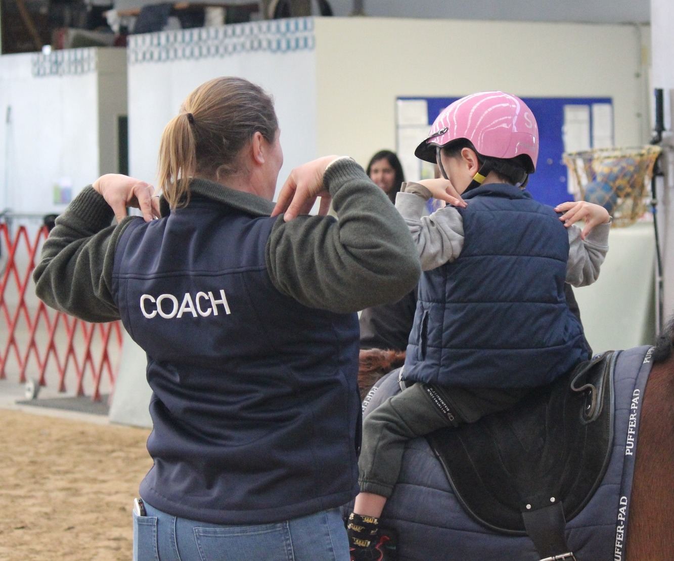 A coach in an RDA vest helping a young rider in a pink helmet stretch while sitting on a horse
