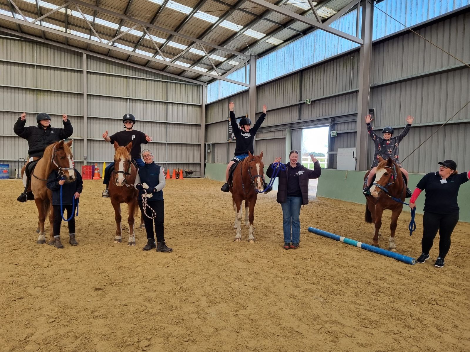 Four riders on horseback raising their arms in the indoor arena while volunteers lead and coaches watch