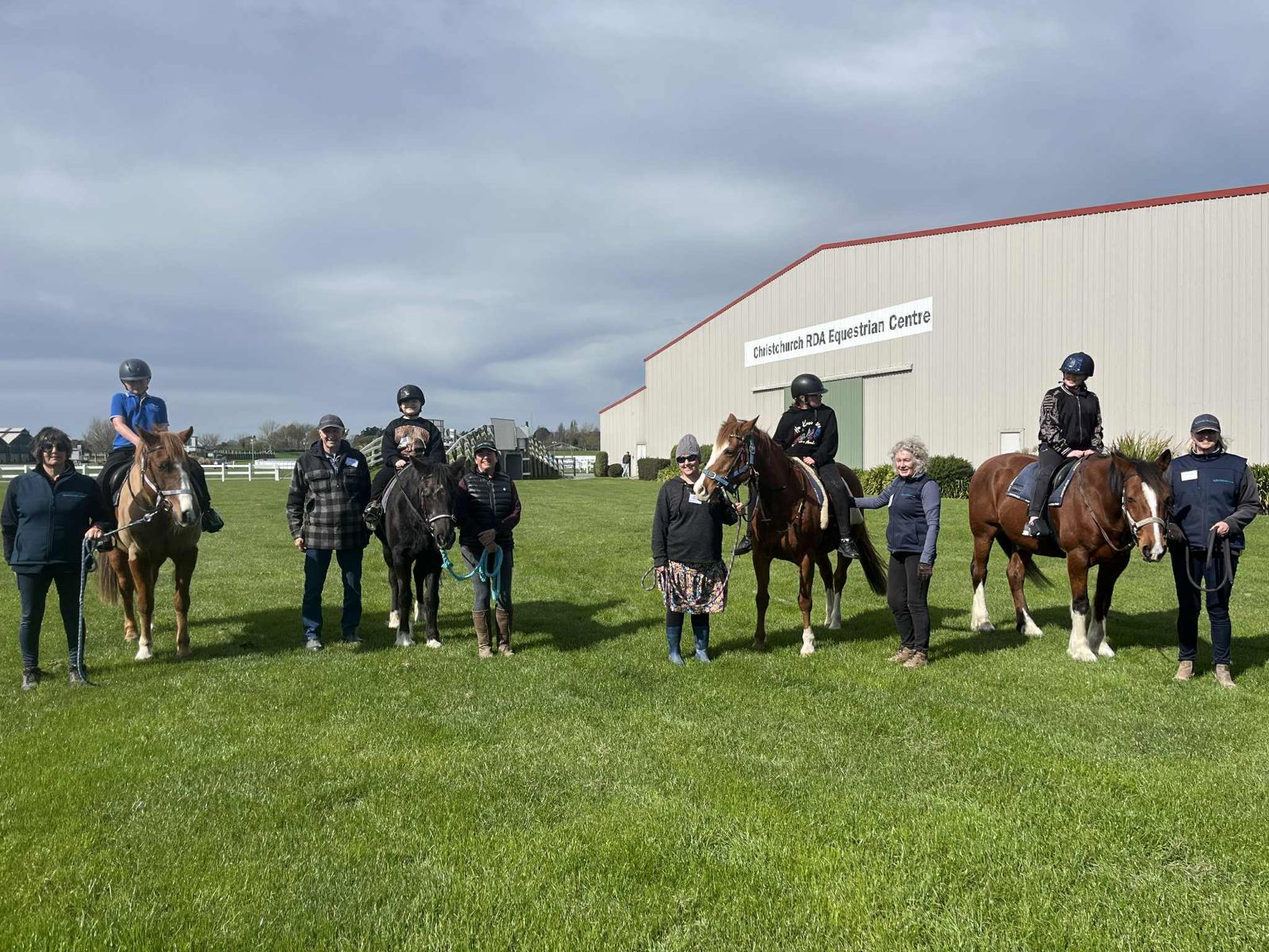 Riders on horseback and volunteers lined up on the grass outside the arena building