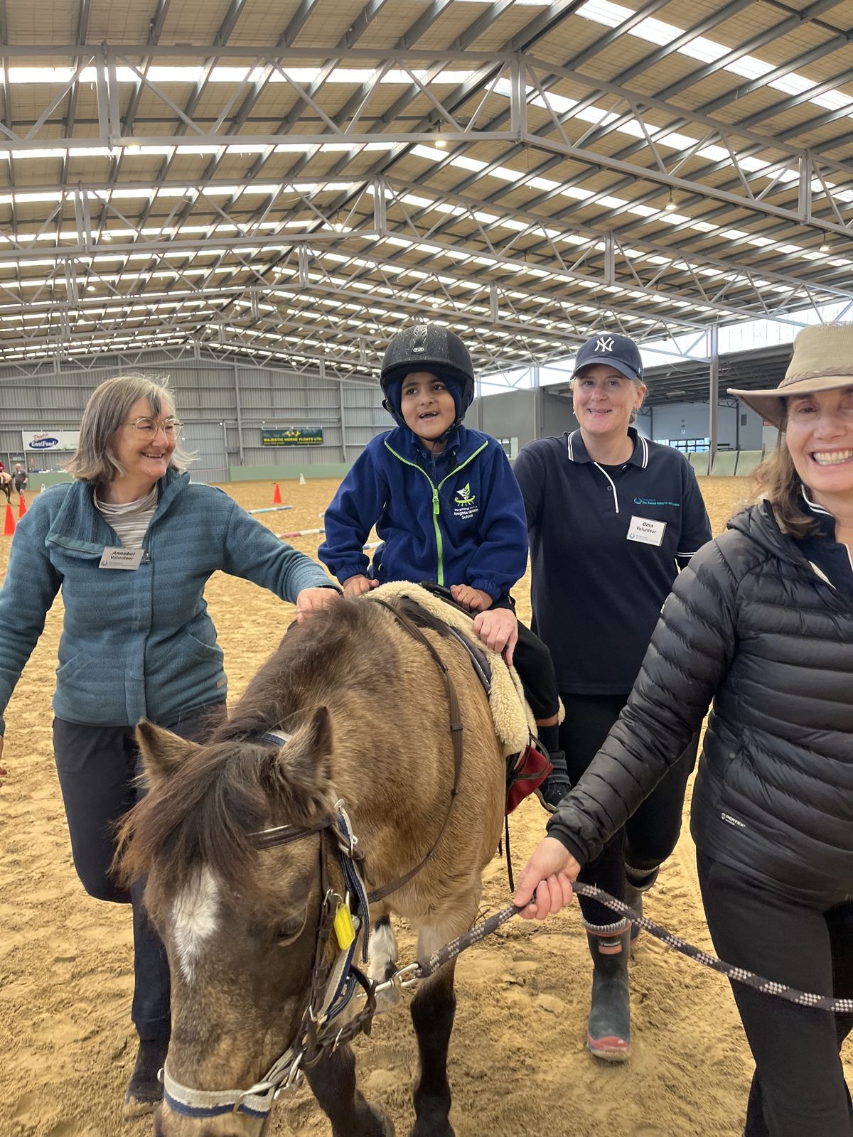 A young rider on a pony, led by a volunteer and supported by side-walkers in the arena