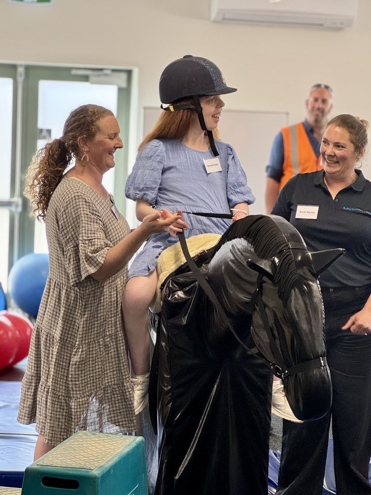 A rider in a helmet sitting on a dark horse, supported by a coach and two volunteers in the therapy room