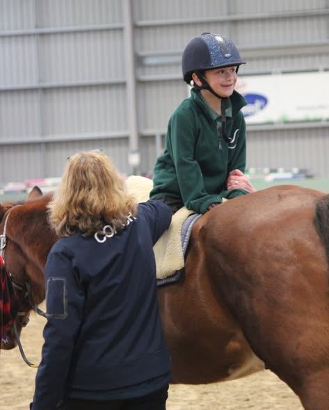 A coach in a navy jacket walks beside a smiling rider in a green jumper on a chestnut horse