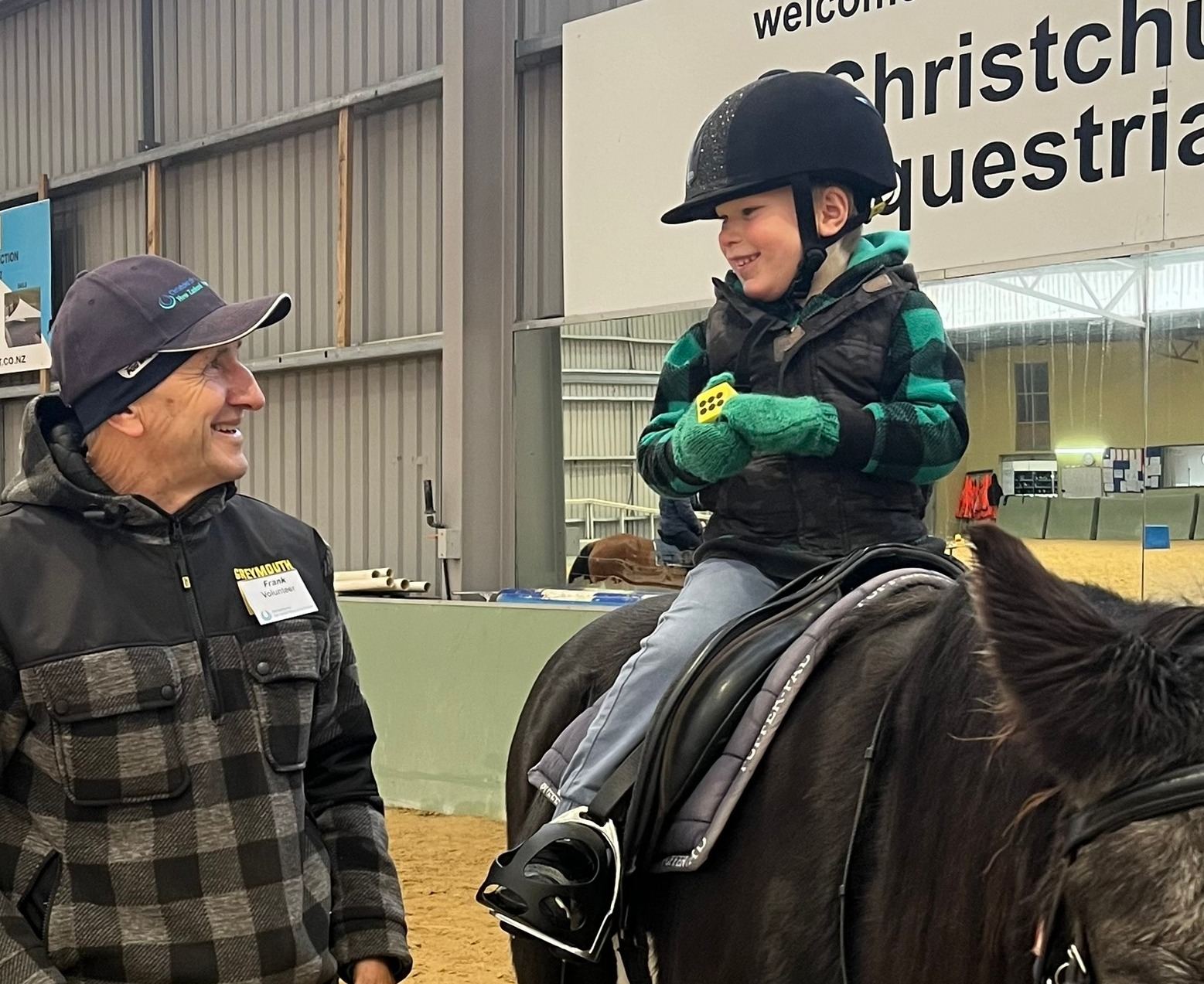 A smiling volunteer in a cap standing beside a young rider in a green jacket on a dark horse